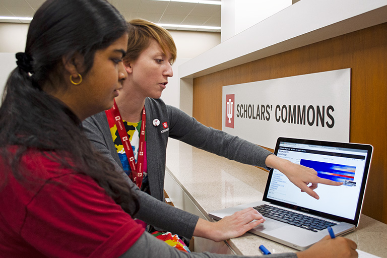 Two woman standing in front of a "Scholars Commons" sign look at a computer; one woman is gesturing toward the screen. 