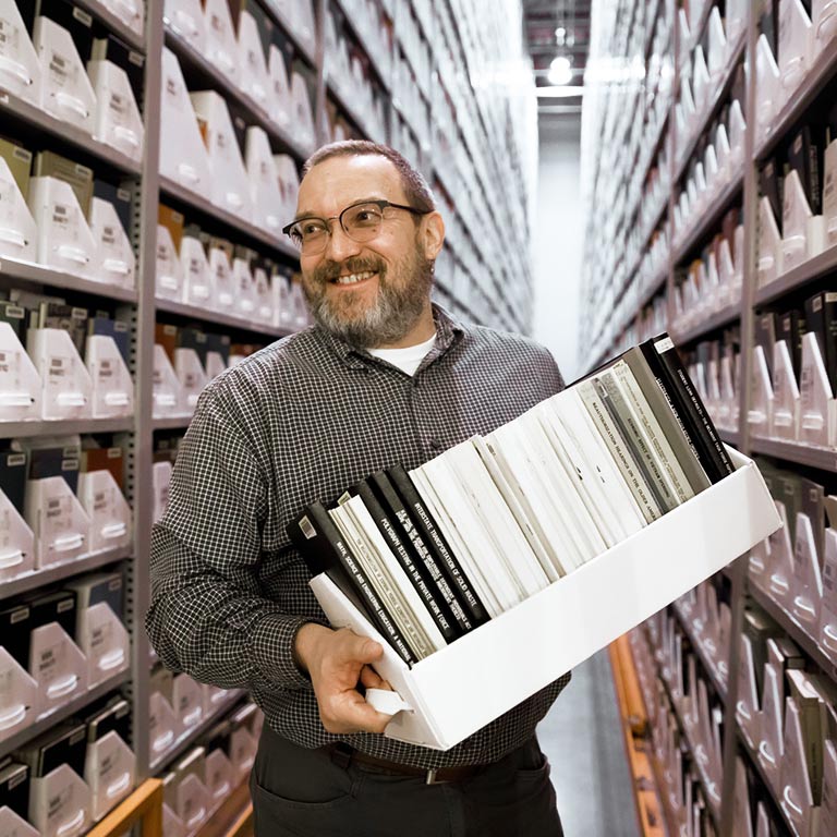 A smiling man carries a box of books between the tall stacks in the Lilly Library. 