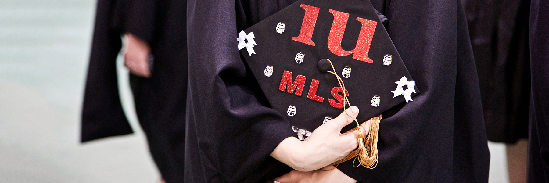 A student wearing a graduation gown shows the top of her graduation cap, which says M.L.S. in red tape.