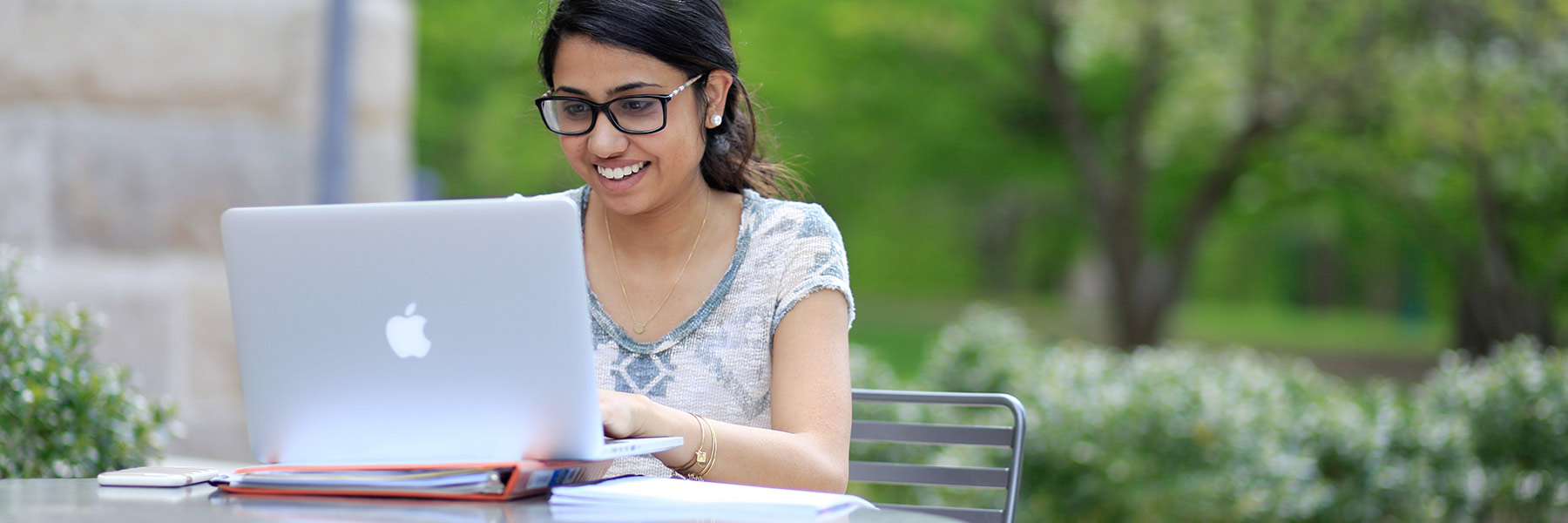 A woman sits outdoors surrounded by green trees while she works on her laptop. 